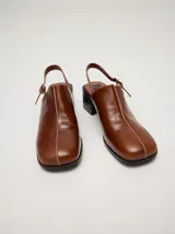 Zuecos de cuero vacuno con acabado en charol, color beige. Presentan un diseño clásico con taco cuadrado de madera de 5 cm y correa trasera ajustable con hebilla metálica.