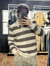 Buzo de tejido de punto a rayas horizontales en tonos beige y marrón, con cuello polo y medio cierre frontal. Presenta puños y ruedo acanalados.