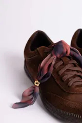 Zapatillas de gamuza color marrón con suela de goma color caramelo. Presentan cordones al tono y un detalle decorativo de pañuelo estampado atado en los laterales con esferas metálicas doradas.