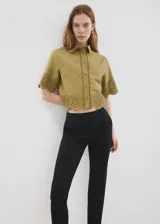 Camisa crop de algodón color verde militar, con cuello camisero, manga corta y cierre frontal con botones. Presenta detalles bordados calados y terminaciones festoneadas en el ruedo y las mangas.