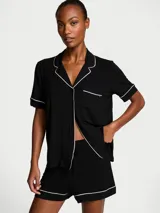 Pijama corto de modal negro compuesto por una camisa de manga corta con botones, bolsillo en el pecho y cuello solapa, y un short a juego con cintura elástica y cordón ajustable. Ambas piezas cuentan con ribetes blancos en contraste.