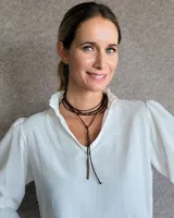 Collar tipo corbatín confeccionado en tiento de cuero marrón, con diseño de doble vuelta y detalle de cuentas metálicas doradas distribuidas a lo largo de las tiras.