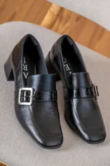 Mocasín de cuero vacuno color negro con acabado texturizado, punta cuadrada, taco bloque y tira decorativa con hebilla metálica plateada en el lateral.