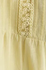 Blusa de tejido liviano color amarillo claro, con escote en V profundo, mangas cortas estilo mariposa y cierre frontal con botones. Presenta detalles de alforzas e inserciones de encaje en el frente y en el borde del ruedo y las mangas, terminando en un volante inferior.