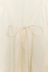 Blusa de color beige con escote en pico y lazo frontal decorado con abalorios. Presenta mangas largas con puños elásticos y un detalle de volantes amplios con terminación de puntilla a tono sobre los hombros y el pecho.