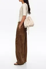 Bolso tipo saco color beige de piel vacuna, con estampado de palmeras pequeñas en marrón, cordón de ajuste frontal y asa regulable con hebilla metálica. Incluye el logo grabado de la marca en la parte inferior.