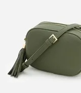 Cartera bandolera pequeña color beige, confeccionada en poliuretano con textura granulada. Presenta cierre superior con cremallera, correa larga ajustable y detalle de borla decorativa en el lateral con herrajes dorados.