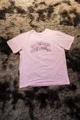 Remera de algodón elastizado color rosa pálido, con cuello redondo y manga corta. Estampado frontal en tonos morados con texto que dice "ARTE INMORTAL CUERPO MORTAL" y un diseño de relámpago.