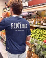 Camiseta de fútbol de Escocia de manga corta, color azul marino con diseño a cuadros sutiles, cuello en V y cuello camisero blanco con líneas azules. Presenta detalles estampados en el frente, incluyendo el escudo de la Asociación de Fútbol de Escocia, un león rampante y el texto 'Scottish Football Association'.