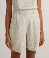 Bermuda corta color beige con estampado floral bordado, de corte sastrero, con bolsillos frontales, cierre por cremallera y botón de presión.