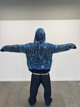 Buzo tipo hoodie tejido de punto grueso con cierre frontal completo hasta la capucha. Presenta un diseño gráfico abstracto en tonos azules y negros, con tipografía estilo gótico en el frente y las mangas. Cuenta con puños y ruedo acanalados en color negro, y etiqueta de marca en la manga.