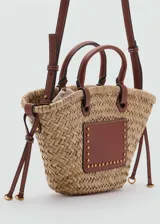 Bolso tipo canasta de paja con aplique de cuero marrón con tachas doradas, doble asa de mano y correa bandolera desmontable.