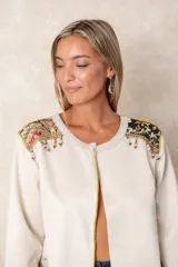 Chaqueta larga color beige con diseño abierto, cuello redondo y mangas largas. Presenta detalles bordados artesanales con aplicaciones de cuentas y toques dorados en la zona de los hombros.