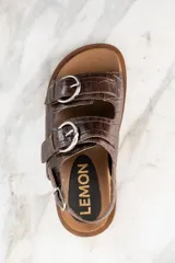 Sandalias bajas color marrón con textura croco, dos tiras anchas sobre el empeine con hebillas metálicas plateadas y tira trasera ajustable con hebilla.