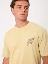 Remera de manga corta color amarillo, corte holgado, con gran estampado en la espalda de un perro en bicicleta con una tabla de surf y un loro, acompañado del texto "Wild by Nature Rusty Worldwide".