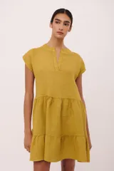 Vestido corto de lino color mostaza, con escote en V y mangas cortas caídas. Presenta un corte holgado con un fruncido a la altura de la cadera que genera un efecto de volados en la falda.