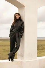 Mameluco negro de cuero ovino, con cuello alto, cierre frontal con botones a presión, cinturón con hebilla forrada y bolsillos laterales.