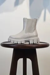 Bota tipo Chelsea de caña corta en color beige, confeccionada en material sintético con acabado liso. Presenta paneles elásticos laterales a tono y una suela track robusta y dentada que aporta altura y estilo moderno.