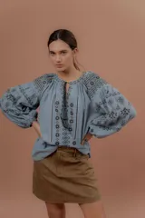 Blusa de estilo bohemio color celeste, con mangas abullonadas y amplias, escote redondo con tiras colgantes y borlas, y detalles de bordados geométricos en contraste de color en el pecho y las mangas.