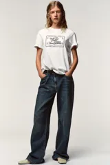 Camiseta blanca de manga corta y cuello redondo, con aplicación de organza y texto negro a contraste en el pecho.