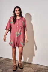 Vestido camisero corto de color rosado con estampado de aves abstractas en tonos verdes y azules. Cuenta con cuello camisero, cierre frontal con botones, mangas cortas y un bolsillo aplicado en el pecho.