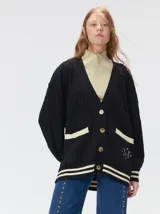 Cardigan de punto trenzado en color negro con detalles en color crema en los bolsillos, puños y ruedo. Presenta un diseño oversize, cierre frontal con botones y bordado decorativo en uno de los bolsillos delanteros.
