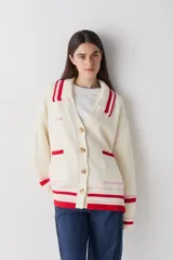 Cardigan de punto color crema con detalles en rojo y rosa en cuello, puños y ruedo. Presenta cierre frontal con botones, cuello tipo polo y dos bolsillos delanteros.