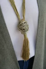 Collar largo de estilo artesanal con cordones de hilo color verde oliva, que presenta un dije central esférico compuesto por hilos metálicos entrelazados y una borla colgante del mismo tono.