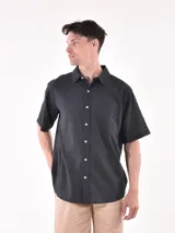 Camisa de manga corta color negro, con cuello camisero y botones blancos frontales. Presenta un bolsillo de parche en el pecho izquierdo y un corte holgado.