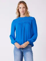 Blusa azul de gasa con pliegues en la parte delantera y detalles de encaje calado. Cuello alto fruncido y mangas largas con puños elastizados. Espalda lisa con abertura tipo gota y botón.