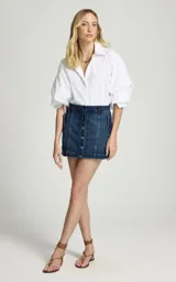 Short pollera de jean de denim azul, de corte recto y tiro alto, con cierre frontal de botones metálicos visibles, trabillas para cinturón y costuras verticales decorativas en el frente.