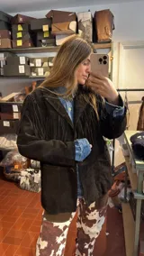 Campera de gamuza negra con corte recto y hombros estructurados. Presenta flecos largos en la parte superior delantera, espalda y mangas, con cierre frontal de botones y bolsillos laterales.