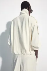 Chaqueta oversize negra confeccionada en hilatura con mezcla de modal, de corte fluido. Cuenta con cuello subido, manga larga con puños elásticos, bolsillos de vivo en el delantero y trabillas en los hombros. El bajo es abullonado con elástico y el cierre frontal es con botones a presión.