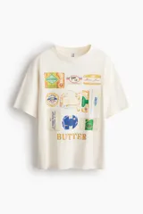 Remera oversized de punto suave de algodón, color blanco, con cuello redondo acanalado, hombros caídos y bajo recto. Presenta un diseño estampado al frente con ilustraciones de diferentes marcas y envases de manteca.