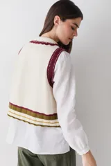 Chaleco de lana color marrón con cuello estilo bebé, cierre frontal con botones grandes y detalle de puntilla en cuello, sisas y bolsillo frontal. Presenta una franja decorativa en color rojo en el ruedo.