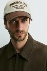 Gorra de seis paneles de algodón en color beige con visera marrón contrastante. Cuenta con un diseño bordado en marrón en el frente con texto alusivo.