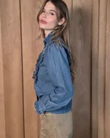 Camisa de denim celeste con cuello alto con volados, botonadura frontal y detalles de volados verticales a lo largo del frente. Cuenta con mangas largas abullonadas y puños con volados.