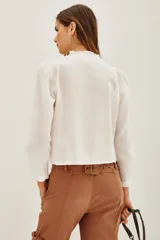 Camisa blanca de algodón con cuello alto, cierre frontal de botones ocultos y detalles de bordado floral en el pecho. Presenta mangas largas con puños elásticos y volados decorativos en la parte frontal.