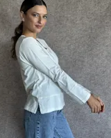 Remera blanca de manga larga con cuello en V y costuras decorativas en contraste. Incluye un lazo ajustable en el cuello con herraje metálico circular.