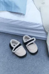 Pantuflas tipo mocasín de corderito color beige con suela y tira de ajuste de velcro en color negro.