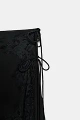 Falda midi negra de tiro medio, confeccionada en tejido satinado, con detalle de aplicación de encaje a tono en el borde lateral y bajo asimétrico. Posee lazadas ajustables en el lateral y cierre lateral oculto.