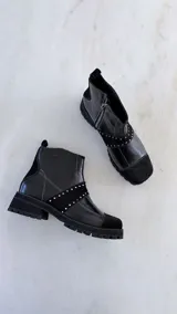 Botín de cuero negro con diseño de paneles combinados en cuero liso y gamuza. Presenta una banda diagonal con tachas metálicas plateadas, costuras decorativas en zigzag y suela track robusta.
