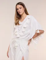 Blusa blanca de silueta holgada con sutil trama en hilo metalizado lurex, escote en V con pechera tejida en crochet y apliques de borlas en el contorno del cuello y las mangas.