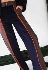 Pantalón de paño con silueta recta y levemente acampanada. Presenta un diseño de paneles combinados en azul marino y marrón, con detalles de vivos en cuero color naranja que recorren el largo de las piernas. Cuenta con cierre frontal y bolsillos laterales.