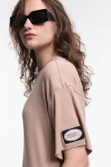 Vestido midi color beige, de corte oversized, con cuello redondo, manga corta y tajos laterales. Presenta un parche decorativo en la manga.