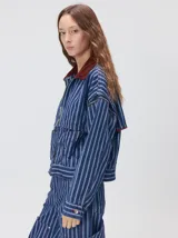 Campera de denim a rayas verticales con cuello de pana en color bordó. Presenta cierre frontal, mangas desmontables con cierre en los hombros, canesú en el pecho y bolsillos de parche cuadrados.