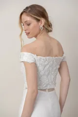 Vestido largo de novia de tul color blanco, con diseño de hombros descubiertos, mangas cortas caídas y corsé con apliques bordados a mano. Presenta una falda sirena con detalles florales bordados y cola.