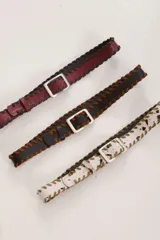 Cinto de cuero con costuras de tientos en los bordes y hebilla metálica cuadrada. Se presenta en tres variantes: cuero bordó con tientos azul marino, cuero marrón oscuro con tientos marrón claro, y cuero blanco con manchas marrones (tipo vaca) con tientos marrón claro.