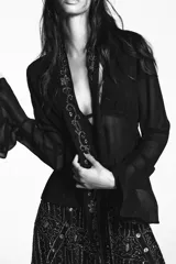 Camisa semitransparente negra de viscosa, con escote en pico pronunciado y manga larga con volantes. Incluye un foulard del mismo tejido con bordados florales y abalorios a contraste en tonos dorados y plateados.