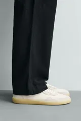 Zapatilla deportiva tipo barefoot confeccionada en piel color blanco roto con apliques de gamuza en tonos grises. Posee puntera redondeada y ancha, cierre mediante cordones al tono y suela de goma flexible en color caramelo con tecnología drop 0.